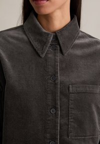 Koszula sztruksowa na guziki w ciemnoszarym kolorze, z kołnierzykiem na spiczasto, delikatną prążkowaną fakturą oraz kieszenią na piersi z dopasowanymi ciemnymi guzikami.