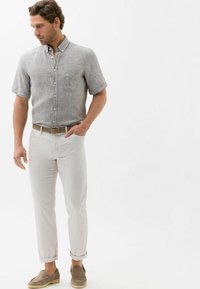 Grijs geblokt overhemd met korte mouwen, witte slim-fit broek, lichtbruine textuurbelt, beige instappers, ontspannen houding tegen een effen achtergrond.