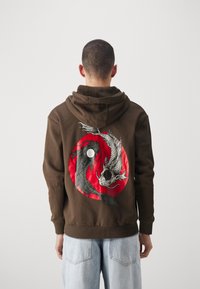 Uomo di spalle che indossa una felpa marrone con pesci koi bianchi e neri che si circondano all'interno di un cerchio rosso sul retro, abbinata a jeans azzurri.