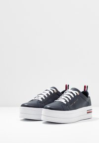Une paire de baskets plateformes bleu marine avec des lacets blancs, des semelles blanches épaisses et des languettes de talon rayées rouge, blanche et bleue ainsi que des accents de semelle.