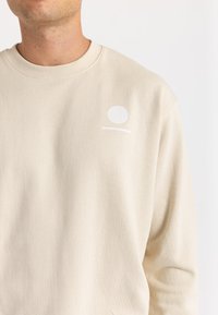 Beige sweatshirt with a round white logo and horizontal line accent near the neckline. Made from soft, textured fabric, with a crew neck design.
