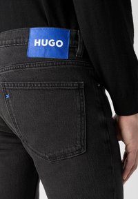 Black denim jeans with a blue leather patch labeled "HUGO" on the back waistband, featuring a single blue stitching detail on the pocket.