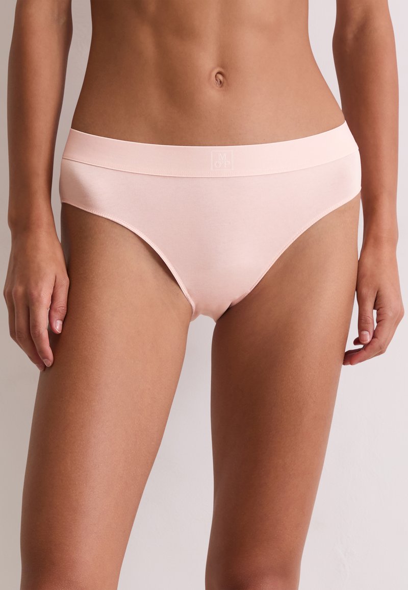 Kobieta nosząca jasnoróżowe bawełniane figi hipster z gładką tasiemką w pasie, stojąca na tle jednolitej, jasnej ściany.