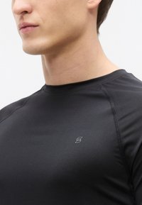 Svart atletisk tröja med korta ärmar, slät textur och en diskret logotyp på vänster bröst. Har rundad halsringning och minimala sömmar.