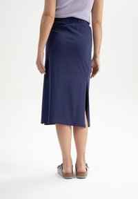 Marineblauwe, knie-lange rok van zachte stof met een elastiche taille en een zijsplit, gecombineerd met grijze sandalen aan de voeten met zichtbare tatoeages.