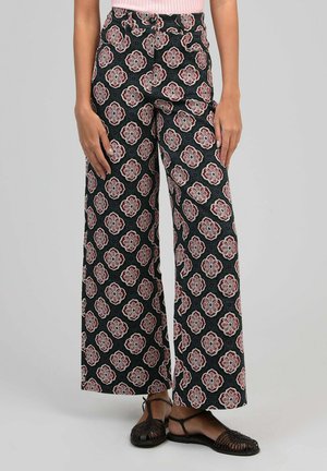 Pantalon noir à jambes larges avec motif géométrique floral rose et blanc, porté avec des sandales en tressé noir et un haut rose clair.