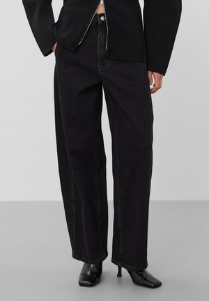 Personne portant un jean noir taille haute à jambes larges assorti à des bottines noires à talons et bouts pointus sur un fond uni.