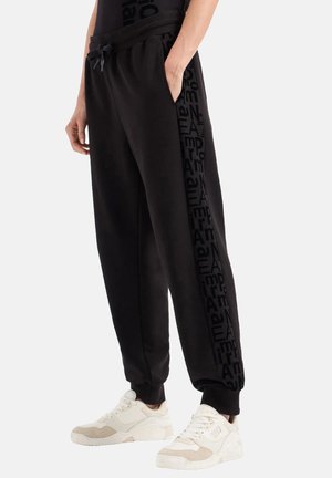 Pantaloni jogger neri realizzati in materiale morbido, con cintura elastica, tasche laterali e una stampa logo tonale ripetitiva lungo la gamba.