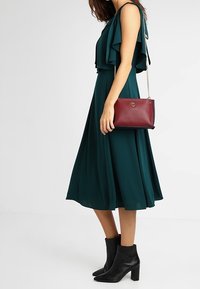Vestido midi de color verde azulado con mangas acampanadas, combinado con un bolso cruzado color granate con cadena. Botines negros completan el conjunto.
