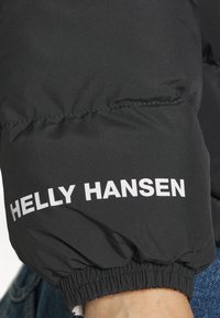 Black puffer jacket sleeve, featuring a white "HELly HANSEN" logo and elastic cuff. Smooth fabric with a matte finish and puffed texture.