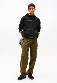 Sweatshirt preta com capuz e logo "TOMMY JEANS", calças cargo verde oliva e sapatos pretos. Detalhes notáveis incluem um boné cinza e as bainhas das calças dobradas.