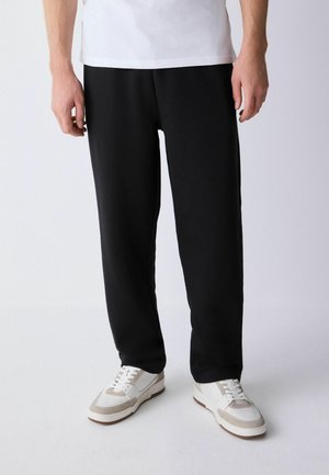 Mann trägt schwarze, gerade geschnittene Hose, weiße Turnschuhe und ein weißes Hemd, steht vor einem schlichten hellen Hintergrund.