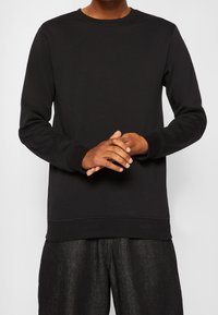 Felpa nera a girocollo realizzata in tessuto morbido, con maniche lunghe e polsini a coste. Indossata con pantaloni scuri e texture.