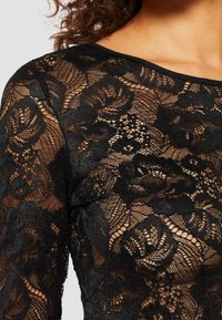 Black lace top featuring a floral pattern, sheer texture, and a classic neckline, with intricate detailing throughout the fabric.