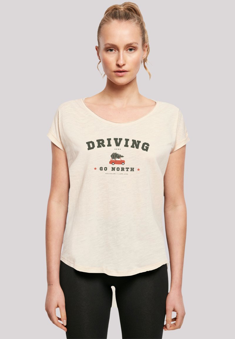 Modell trägt ein hellcremefarbenes T-Shirt mit dem Aufdruck "DRIVING GO NORTH" und einer Illustration eines Autos, das einen Baum transportiert, kombiniert mit schwarzen Leggings.