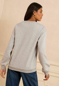 Siv sweatshirt s krojnim izrezom, rebrastimi manžetami in robom ter mehkim teksturo. V kombinaciji s temno modrimi kavbojkami. Prikazano je zadnje gledanje.
