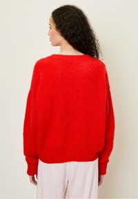 Raudonas pūkuotas megztinis su laisvu siluetu, ilgomis rankovėmis, raukšlėta apatine dalimi ir rankogaliais, turintis plačią apykaklę ir švelnų tekstūrą.