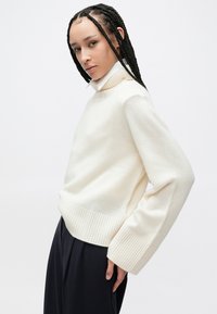 Πλεκτό πουλόβερ σε κρεμ απόχρωση με ριμπ τελείωμα και φαρδιά μανίκια. Διαθέτει ψηλό λαιμό με στρωμένο σχέδιο, φορεμένο πάνω από σκούρο παντελόνι.