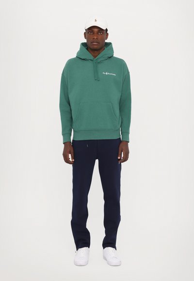 Rohelise kapuutsiga sweatshirt, millel on känguru taskud, koos tumesiniste dressipükste ja valgete spordijalatsitega. Valge müts, millel on logo, täiendavad riietust.