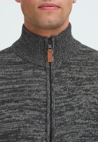 Hombre que lleva un suéter grueso de punto de color gris oscuro con un cuello alto acanalado y un tirador de cremallera de cuero.