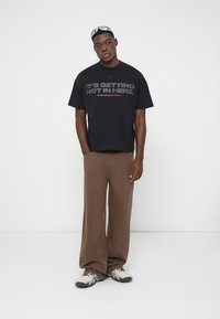 Νέος άνδρας με μαύρη γραφική μπλούζα, καφέ φαρδιά παντελόνια, μπεζ αθλητικά παπούτσια και γυαλιά ηλίου στο κεφάλι, που στέκεται μπροστά από λευκό φόντο.