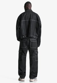 Sort denimjakke og cargo-bukser vist fra bagsiden, med kontrastsyninger og flere lommer, i en afslappet pasform.