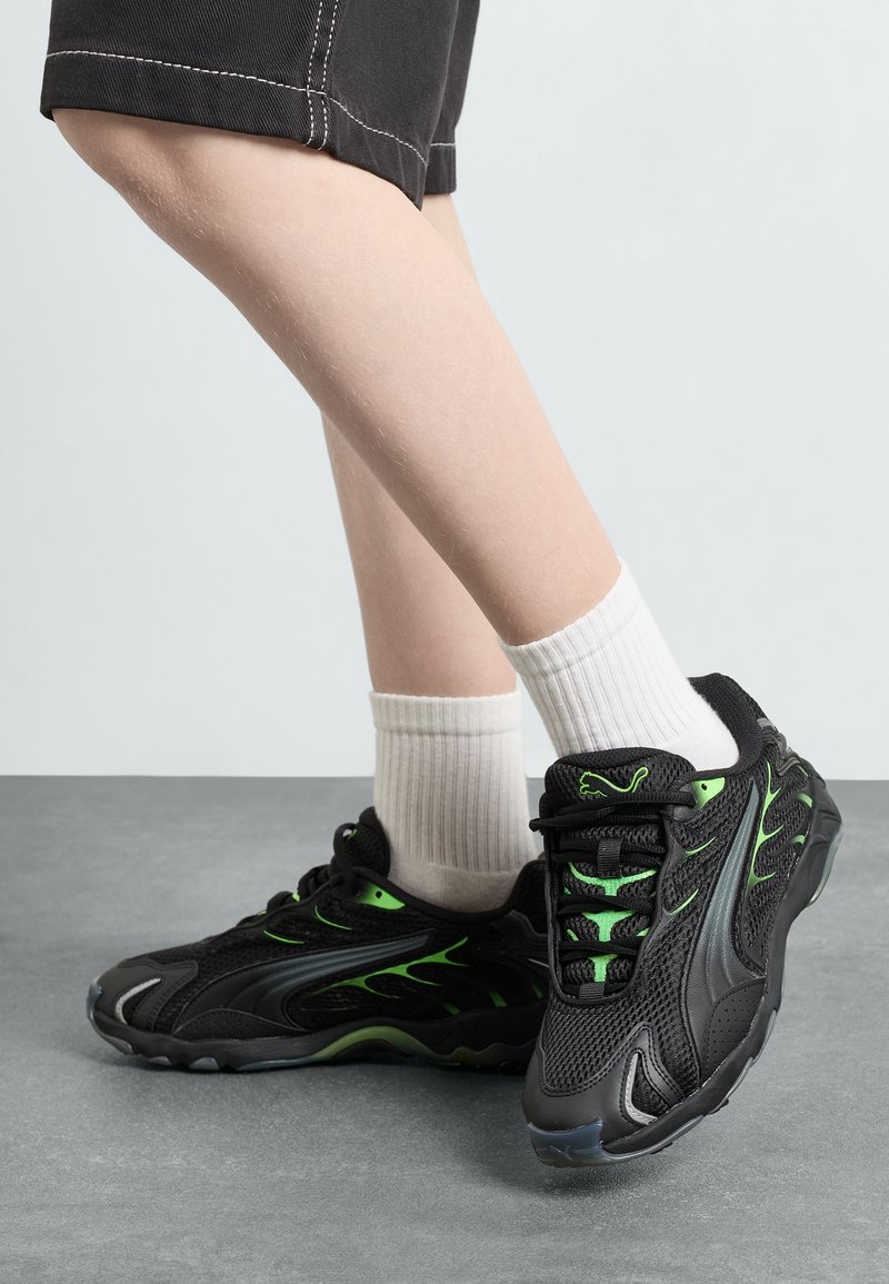 Jambes portant un short noir mi-long, des chaussettes blanches côtelées et des baskets noires avec des détails de flammes vertes et des semelles épaisses.