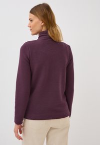 Purple long-sleeve turtleneck top with a textured fabric, featuring a fitted collar and ribbed design, paired with light-colored pants.
