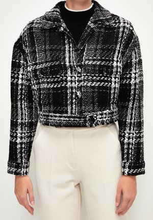 Czarna i biała kraciasta przycięta kurtka z kołnierzem i guzikami noszona na czarnym topie i beżowych spodniach.