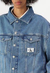 Traper jakna u svijetloplavoj boji, s klasičnim dizajnom i srebrnim dugmadi te CK Jeans logom na džepu na prsima.