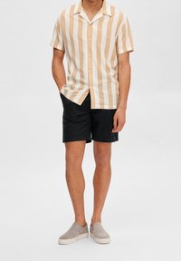 Camicia a maniche corte con righe verticali beige e bianche, abbinata a pantaloni corti neri e scarpe slip-on grigie. Abbigliamento estivo casual.