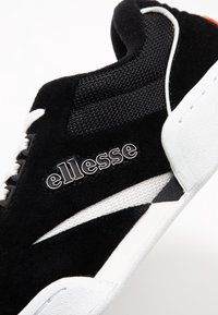 Sneakers en suede noir avec des accents en mesh, une semelle intermédiaire blanche et une semelle extérieure en caoutchouc noir. Le logo "ellesse" est affiché de manière proéminente sur le côté.