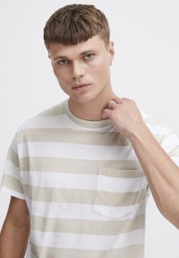 Camiseta de rayas en algodón color beige y blanco, con un bolsillo en el pecho, cuello redondo y mangas cortas. Textura suave y ajuste casual.
