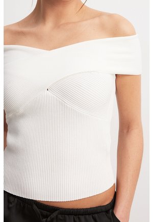 Weißes, geripptes Off-Shoulder-Top mit überkreuztem Ausschnitt, getragen von einer Person, kombiniert mit schwarzen Jogginghosen mit Kordelzug.