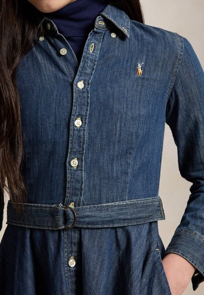 Close-up af en person iført en blå denim knapklædt kjole med matchende bælte og en lille broderet logo på brystet.