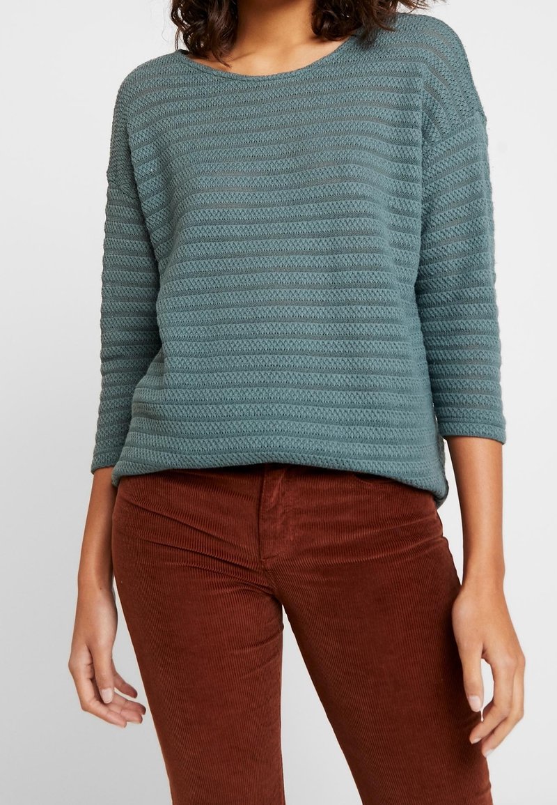 Maglione testurizzato in tonalità teal con righe orizzontali, maniche a tre quarti e scollatura rotonda, indossato sopra pantaloni in velluto a coste color ruggine.