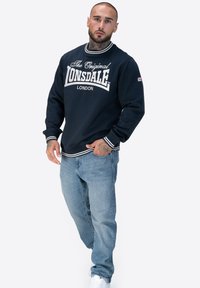 Marine Sweatshirt mit weißem Lonsdale-Logo, geripptem Kragen und Bündchen. Kombiniert mit hellblauen Jeans und weißen Sneakers.