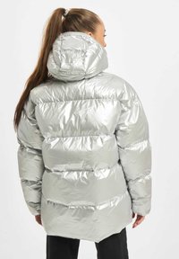 Sølvmetallic oppustelig jakke med hætte, der har en baffle-design, glat tekstur og en forlænget bagkant. Detaljer inkluderer syede sømme.