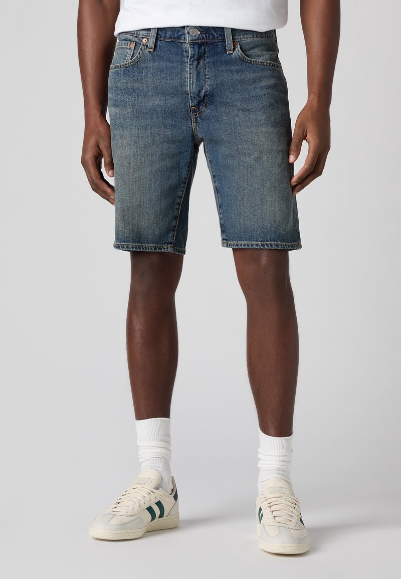 Short en denim bleu moyen avec une coupe droite. Comprend cinq poches, des passants pour ceinture et des détails de couture. Associé à des baskets blanches.