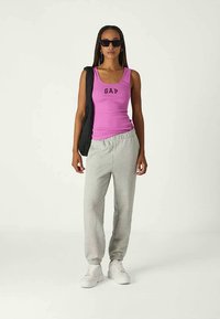 Mulher a usar óculos de sol pretos, top rosa com o logo "GAP", calças de moletom cinzas, ténis brancos e a segurar uma mochila preta.