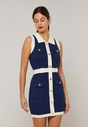 Femme portant une robe sans manches bleu marine avec col blanc, bordure, ceinture, boutons dorés et quatre poches à rabat à l'avant, debout devant un fond uni.