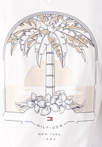 Biely sveter s grafikou palmy v béžovej a čiernej farbe, s kvetmi na spodnej časti a textom "HILFIGER NEW YORK 1985" dole.