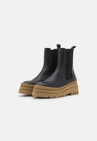 Bottines en cuir noir avec des panneaux élastiques sur les côtés et une semelle en caoutchouc épaisse couleur tan. Présente une texture lisse et un design à bout arrondi.