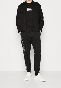 Zwarte overhemd met knopen over een zwart t-shirt, gecombineerd met zwarte trainingsbroek met witte accenten en logo, afgemaakt met witte sneakers.