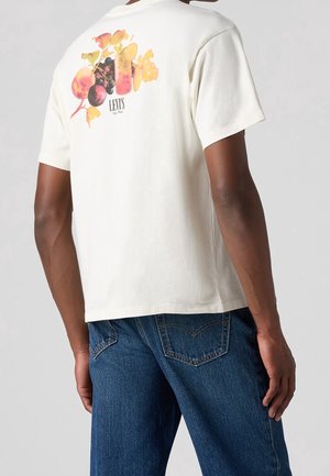 Cremefarvet bomulds-T-shirt med et farverigt frugtprint på ryggen og "Levi's" logoet. Sammen med blå denim jeans.