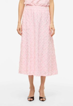 Femme portant une jupe midi en dentelle florale rose taille haute, associée à des talons noirs ouverts, debout devant un fond blanc.