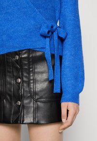 Sininen ribattu neulepaita sivuityllillä yhdistettynä mustaan nahkaiseen napitettuun mini-hameeseen, käsi lepää hameen vieressä.