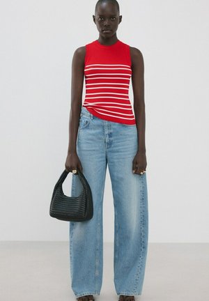 Tânără purtând un top fără mâneci cu dungi roșii și albe, blugi evazați bleu deschis, ținând o geantă neagră țesută, pe un fundal simplu.