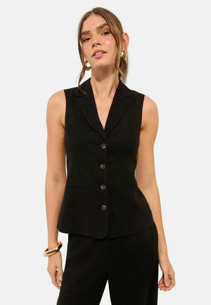 Frau trägt ärmellose schwarze Weste mit Knöpfen und passende schwarze Hose, dazu goldene Creolen und ein massives goldfarbenes Armband.