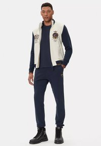 Completo sportivo da uomo composto da un gilet zip grigio chiaro con accenti blu navy e logo, top blu navy e joggers abbinati, accompagnati da stivali neri.